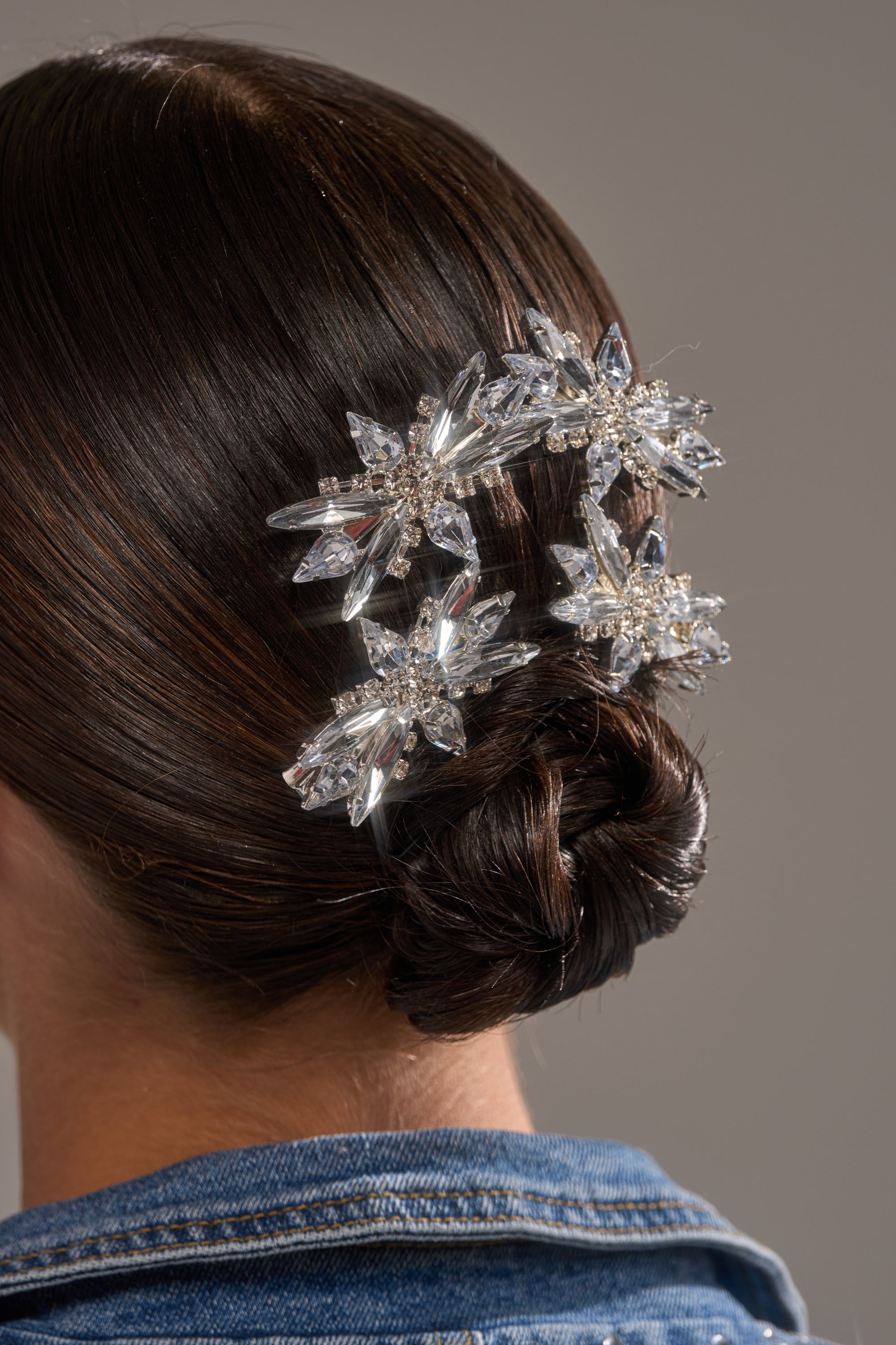 A close-up of a woman’s dark hair in a low bun, styled with two DECO RHINESTONE 4 PK CLIPS shaped like flowers. She wears a blue denim jacket against a plain background. 0