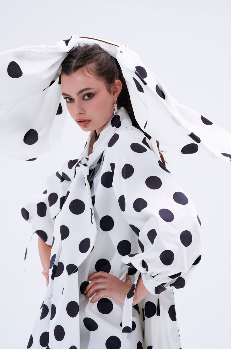 A woman poses confidently in a white dress with large black polka dots and puffed sleeves, accessorized with the POLKA DOT IT HAIR BOW—a matching oversized polka dot headpiece. The background is plain and light-colored.