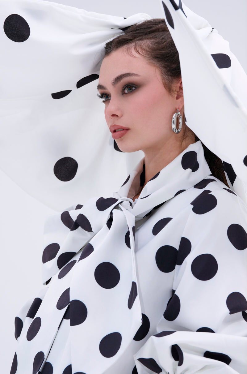 A woman wearing a large white hat with the POLKA DOT IT HAIR BOW, a bold black polka dot coat, silver hoop earrings, and natural makeup stands against a plain background.