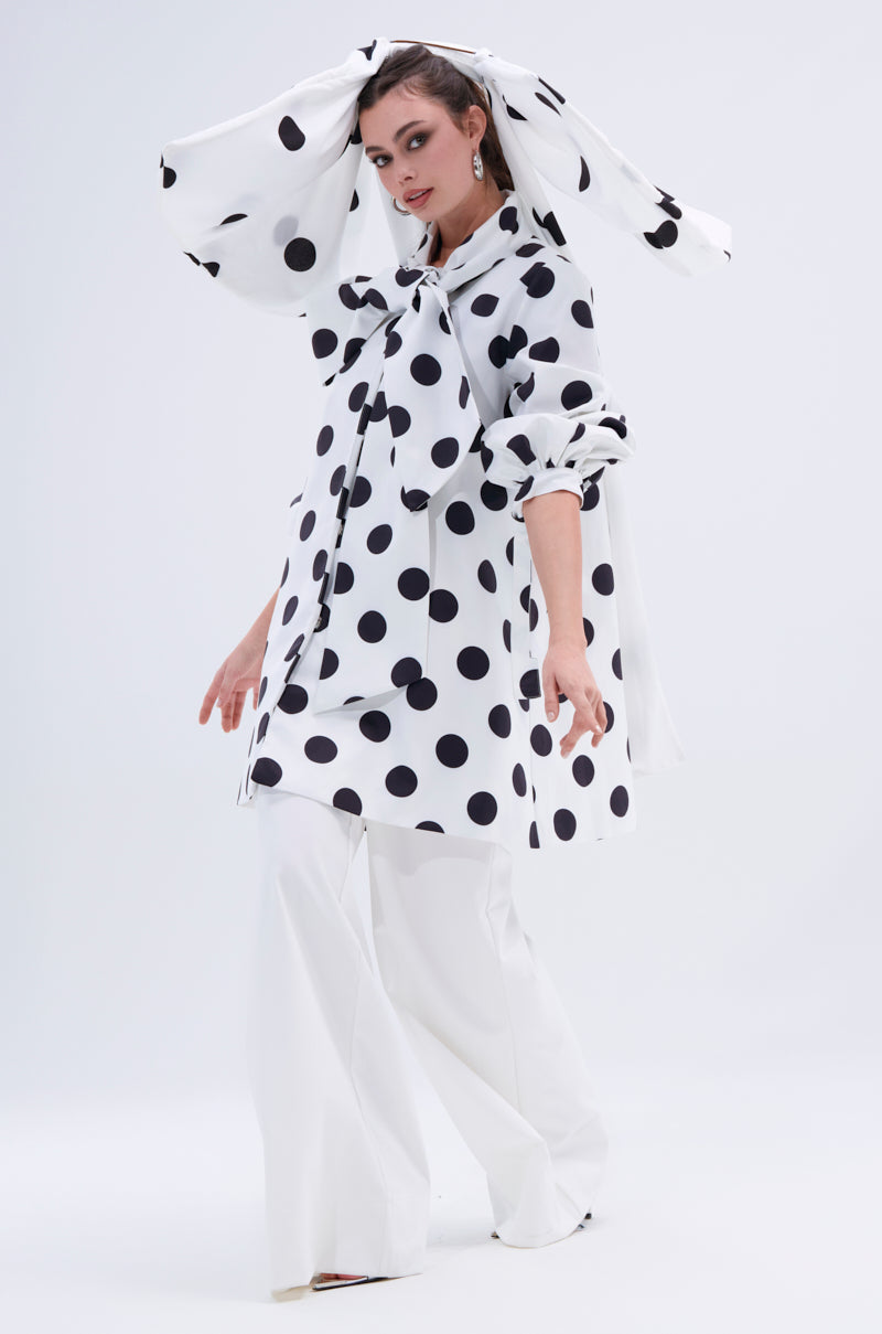 A woman wears wide-legged white pants and a white outfit with large black polka dots, styled with statement earrings and the POLKA DOT IT HAIR BOW, which matches her oversized polka dot neck bow.