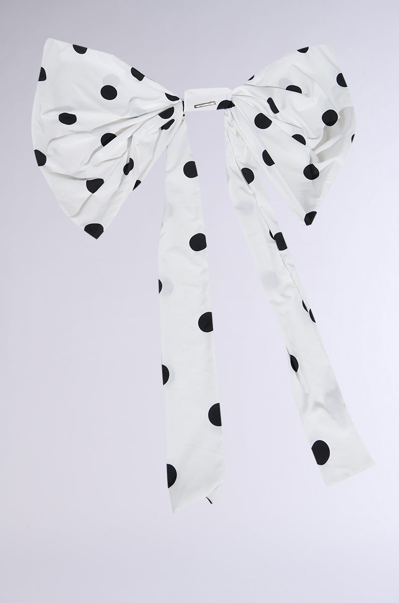 The POLKA DOT IT HAIR BOW is an oversized white hair accessory with bold black polka dots and long trailing ribbons, shown against a plain light background—a statement piece for maximalist style.
