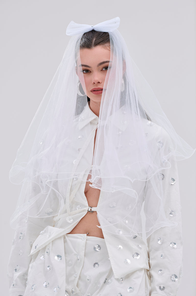 A woman stands against a plain light background, wearing the TO THE EXTREME HAIR BOW atop her head, styled with a sheer white veil and a partially unbuttoned white sequin-and-rhinestone shirt.