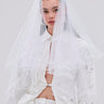 A woman stands against a plain light background, wearing the TO THE EXTREME HAIR BOW atop her head, styled with a sheer white veil and a partially unbuttoned white sequin-and-rhinestone shirt.