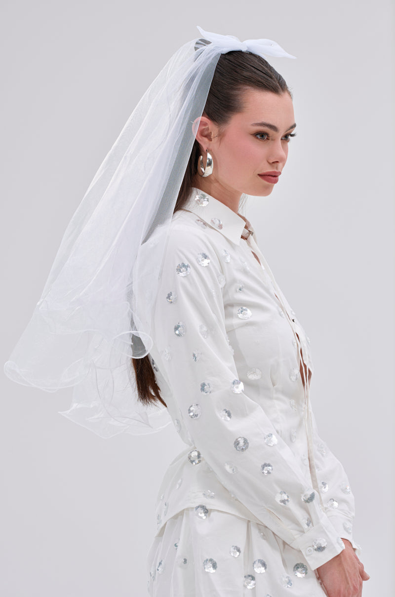 A woman wearing the TO THE EXTREME HAIR BOW, a white outfit with silver sequins, a white veil, and large hoop earrings stands against a neutral background, looking to the side.