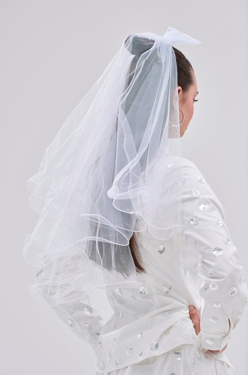 A woman stands with her back to the camera in a white sequin outfit, her hair pulled back and hands on hips, wearing a long tulle veil tied with the TO THE EXTREME HAIR BOW.
