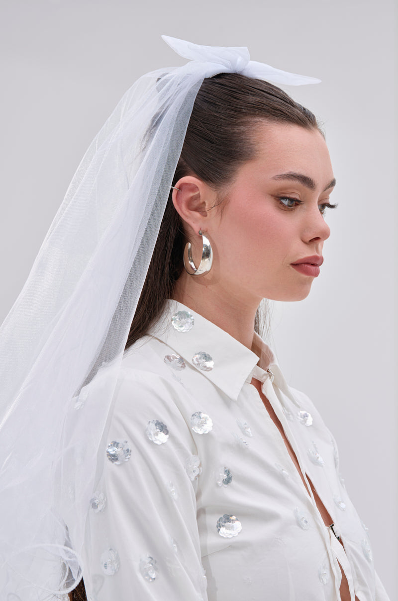 A woman with long brown hair wears the TO THE EXTREME HAIR BOW and a white veil. She pairs this with silver hoop earrings and a rhinestone-detailed white shirt, gazing to the side with a neutral expression.