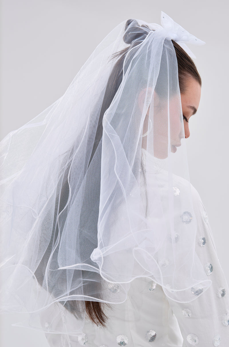 A woman with long brown hair wears the TO THE EXTREME HAIR BOW and a white veil, dressed in a white sequin outfit, looking down against a plain light background.