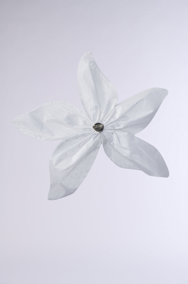 The FLOWER BLOOM BROOCH features a delicate, translucent white flower with five petals and a shiny metallic center, resembling an oversized floral accessory, set against a plain light gray background. 2