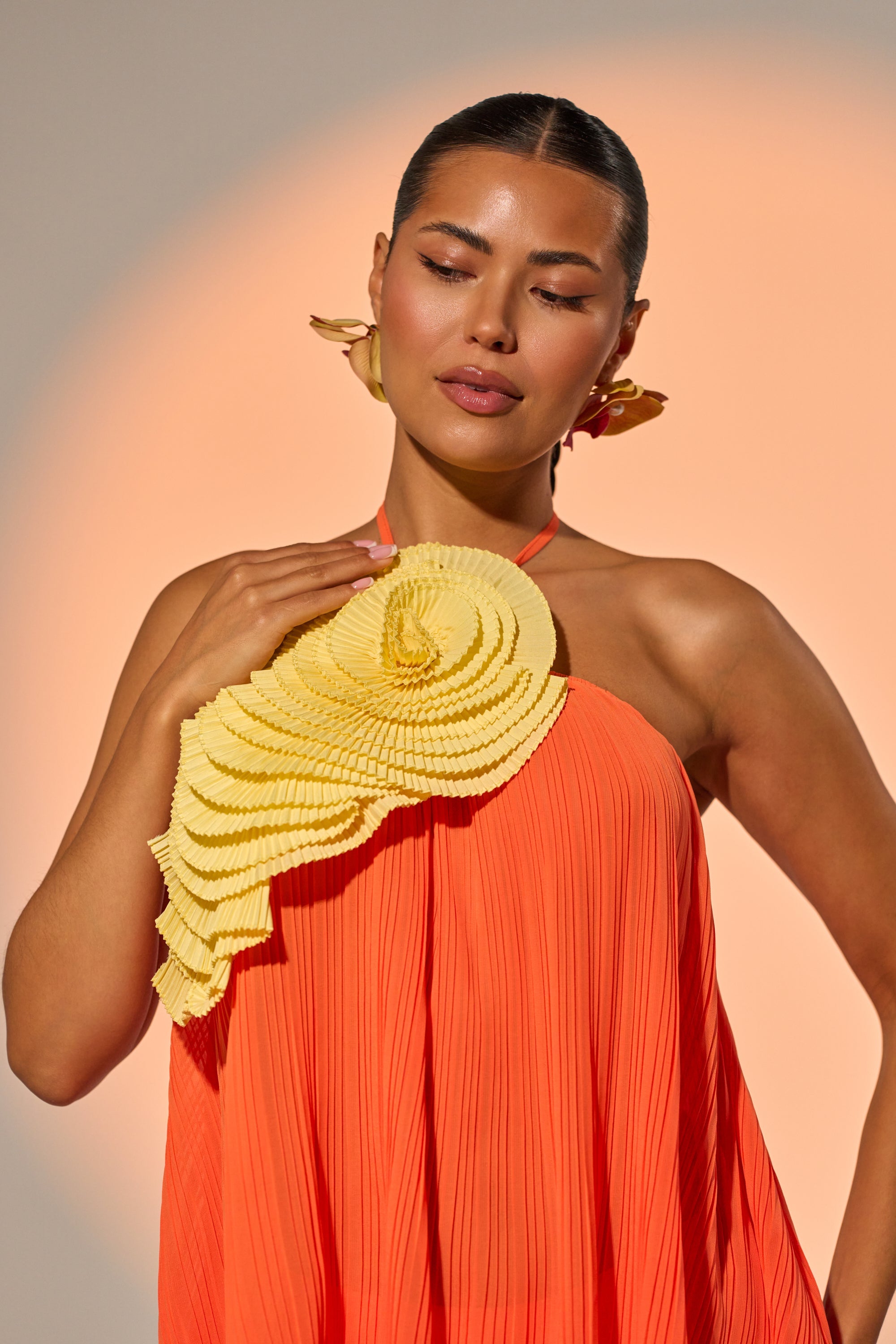 A woman with slicked-back hair poses before a peach gradient, wearing a vibrant pleated orange dress and gold leaf earrings. Her look is highlighted by the IN THE GARDEN OVERSIZED BROOCH and a large, layered yellow chest ruffle. 1