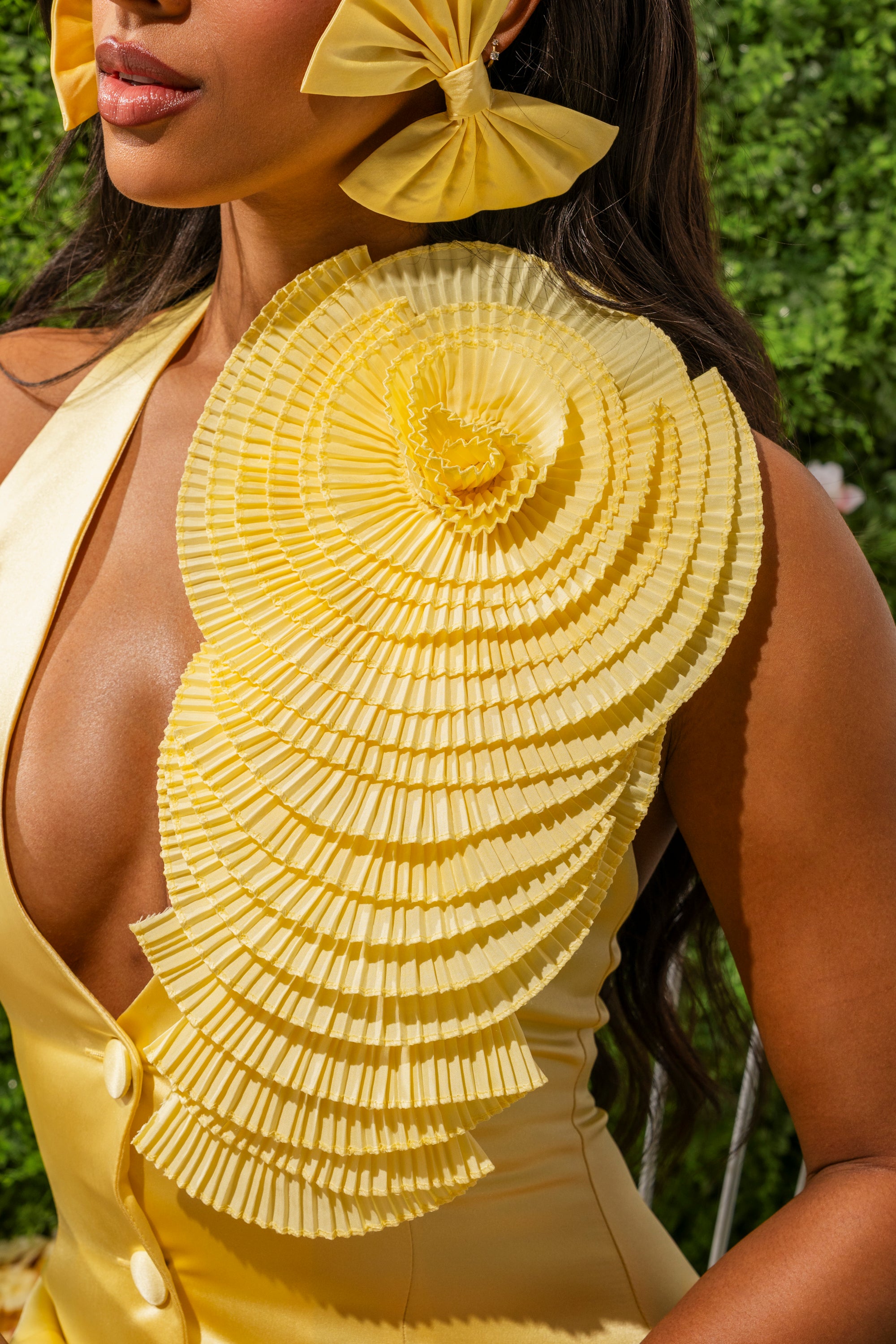 A woman in a sleeveless yellow dress with fan-shaped pleats and a matching hair bow stands outdoors, her look accented by the IN THE GARDEN OVERSIZED BROOCH, a statement accessory amid lush greenery. 0