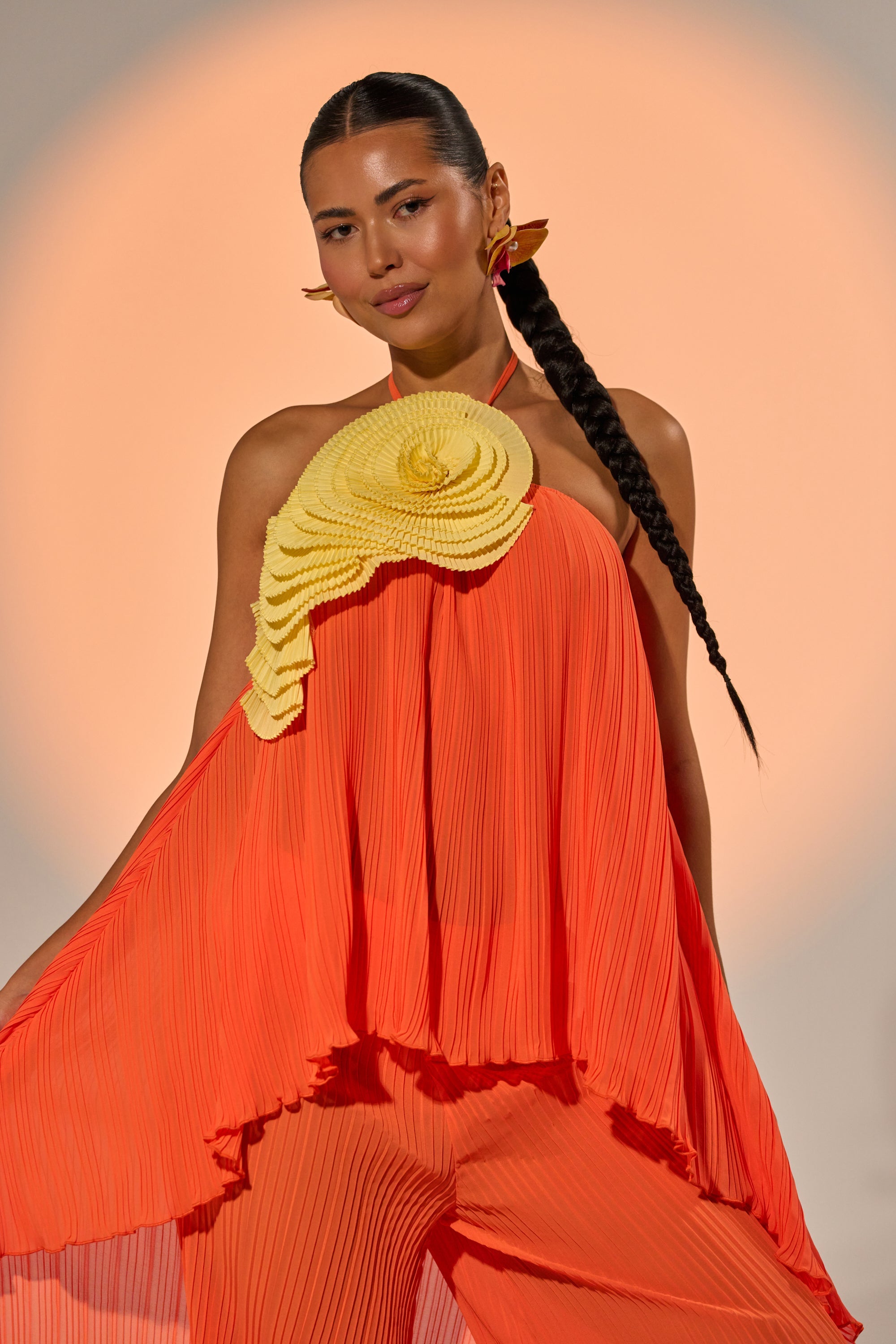 A woman with a long braided ponytail wears a flowing orange dress with layered yellow ruffles and the IN THE GARDEN OVERSIZED BROOCH on her shoulder, posing confidently against a softly lit peach background. 2