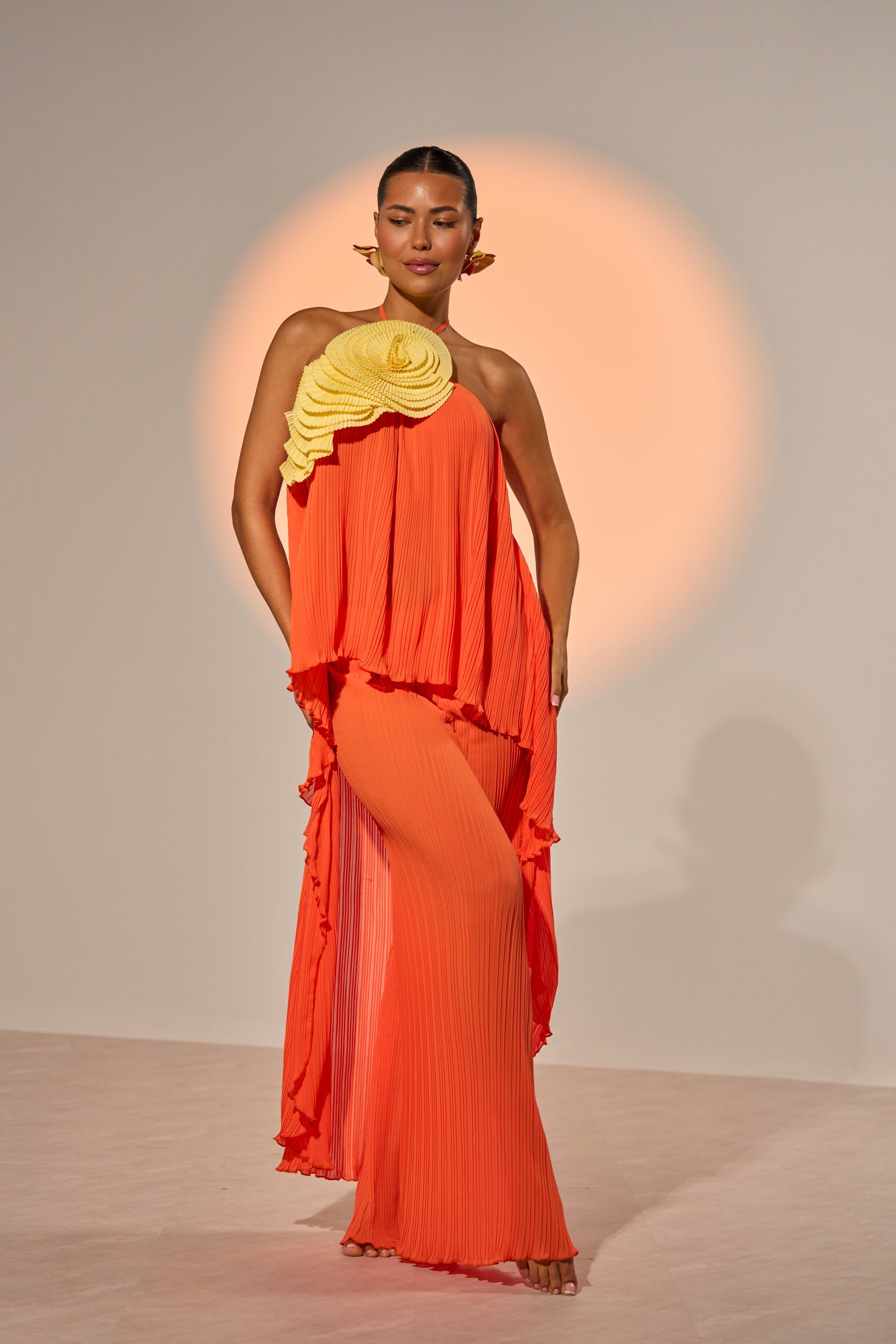 A woman poses in an asymmetrical orange pleated outfit with wide-leg pants and a flowing top, accented by the IN THE GARDEN OVERSIZED BROOCH—a striking rose accessory—set against a neutral background with warm circular light. 3