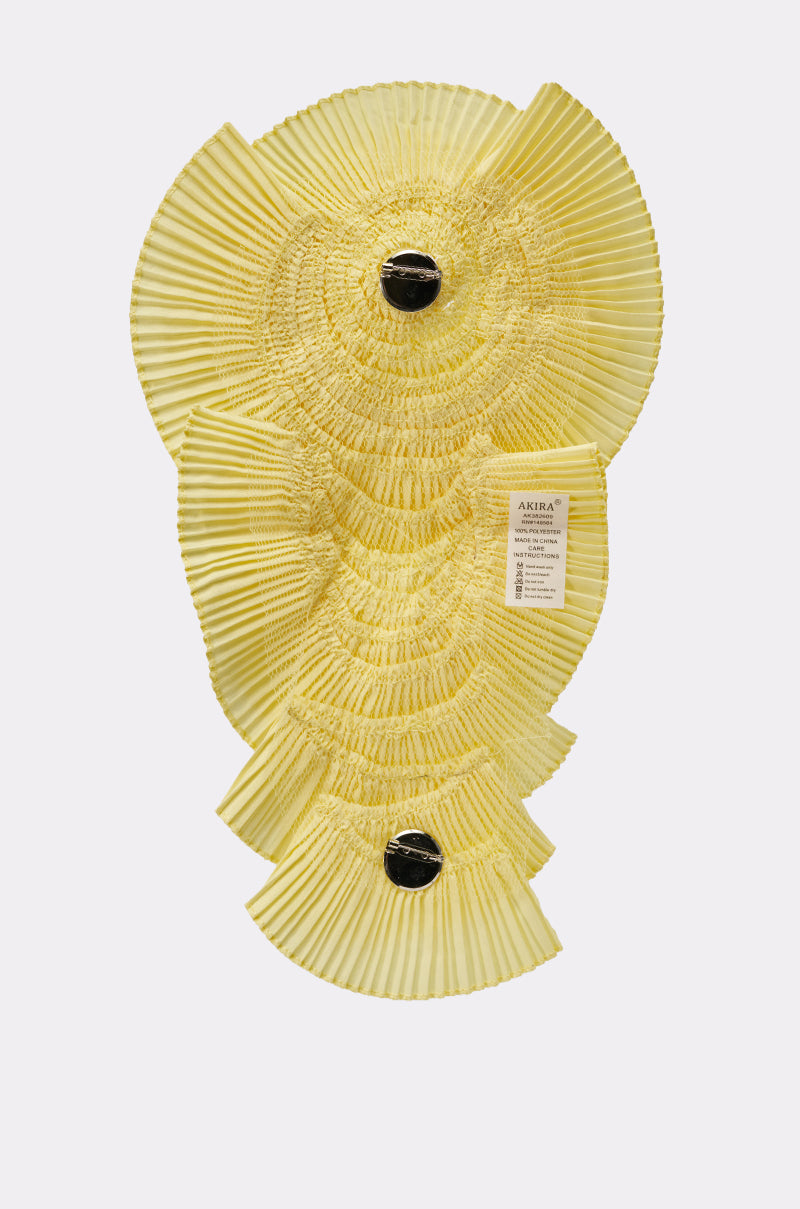 The IN THE GARDEN OVERSIZED BROOCH is a sculptural yellow wall decoration featuring layered, pleated fan shapes, two black circular accents, and a small white tag on the right, displayed on a plain white background. 5