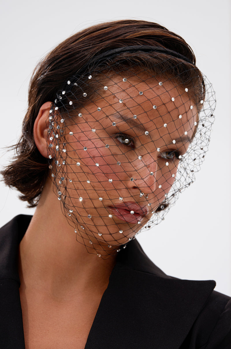 A person with short, dark hair wears the HAILEY BLING HEADBAND, a rhinestone veil accessory covered in sparkling gems. They have natural makeup, a black outfit, and stand against a simple background. 1