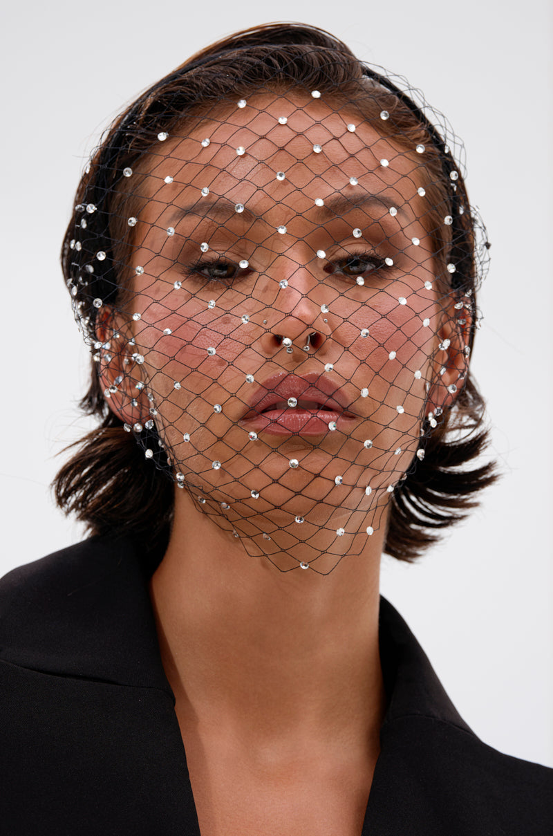 A woman with short dark hair wears the HAILEY BLING HEADBAND, a black netted veil embellished with pearls and silver rhinestones, over a black blazer. She gazes at the camera against a plain white background. 3