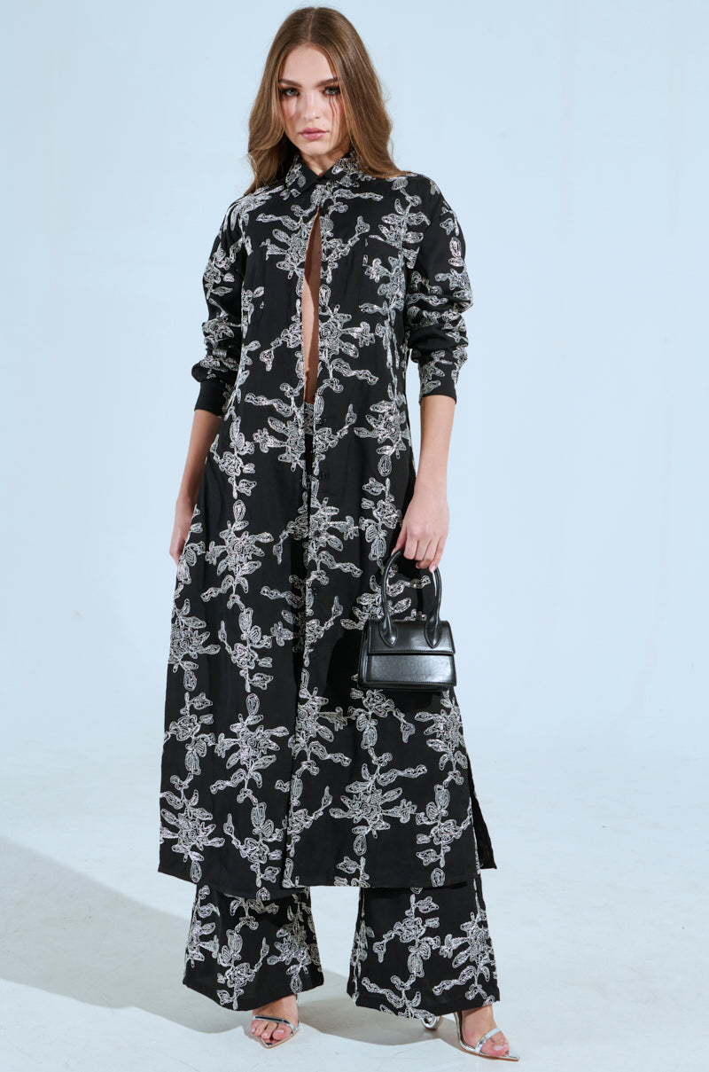 Against a plain light blue background, a person dons the ZIGGY EMBROIDERED LONG BUTTON DOWN TOP. The black coat features intricate white abstract metallic designs, paired with wide-leg pants and a small black handbag.
