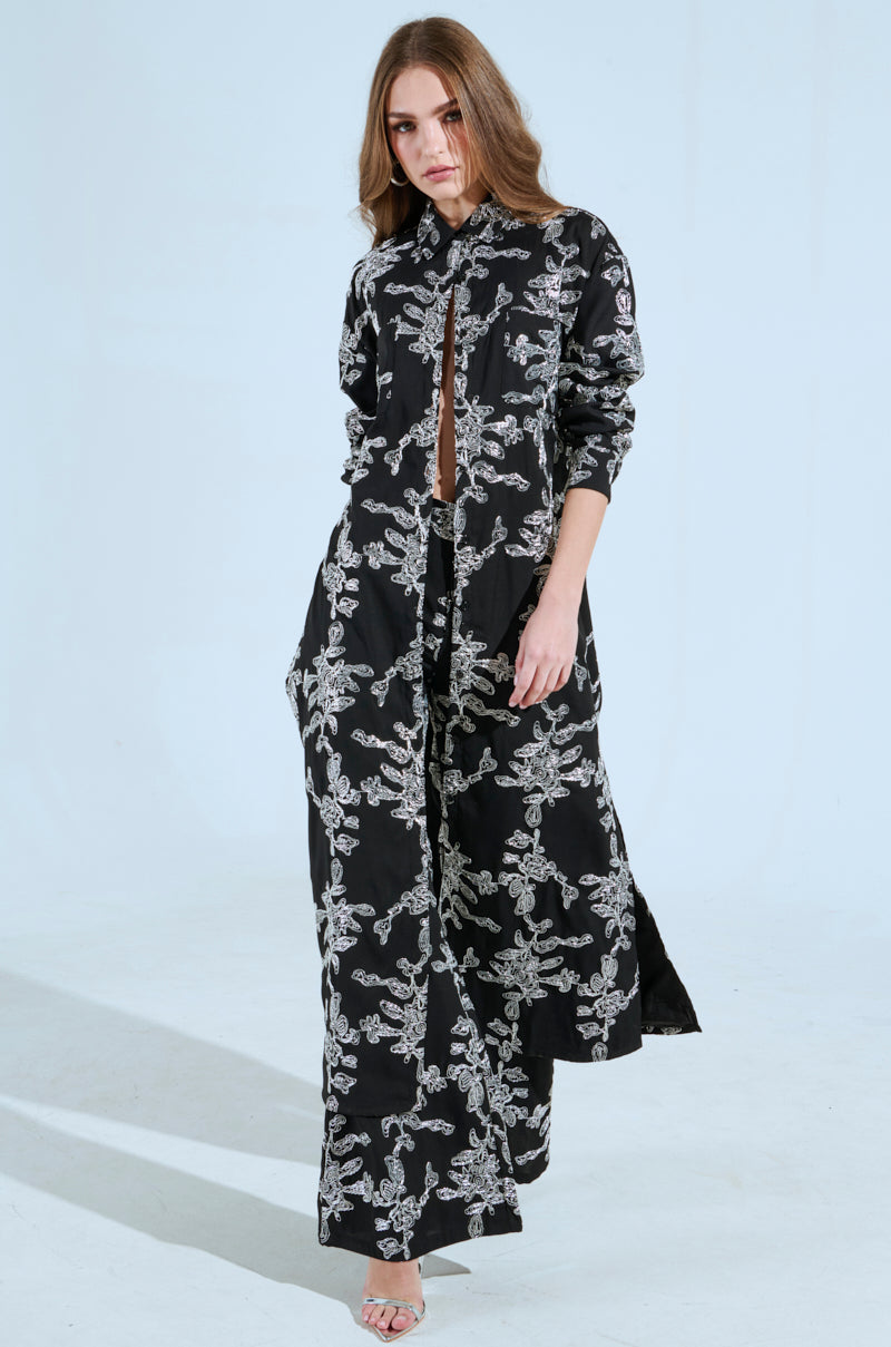 A woman in a ZIGGY EMBROIDERED LONG BUTTON DOWN TOP, with a subtle abstract metallic pattern, stands confidently against a plain background. Her long, wavy hair complements the striking design as she gazes forward, one foot slightly ahead of the other.