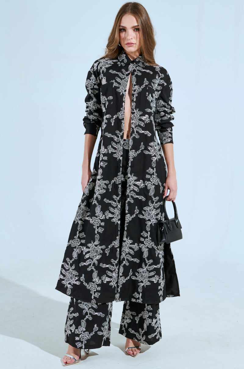 Against a light background, a woman wears the Ziggy Embroidered Long Button Down Top with intricate abstract metallic patterns. She complements her look with a small black handbag and silver open-toe shoes, while her long hair cascades over her shoulders, enhancing the ensemble's elegance.