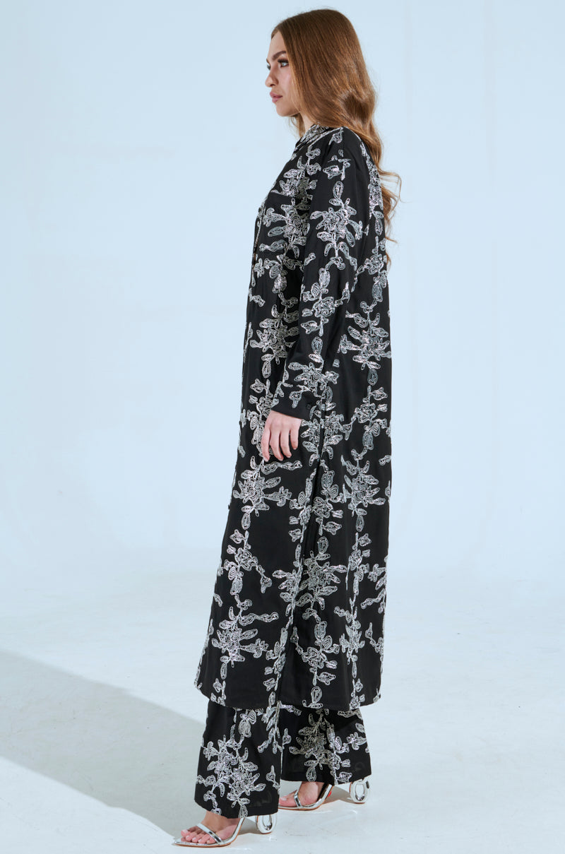 A long-haired woman stands in profile against a plain background, dressed in a ZIGGY EMBROIDERED LONG BUTTON DOWN TOP with an abstract metallic pattern. Her black long-sleeve attire is paired with silver high-heeled shoes.