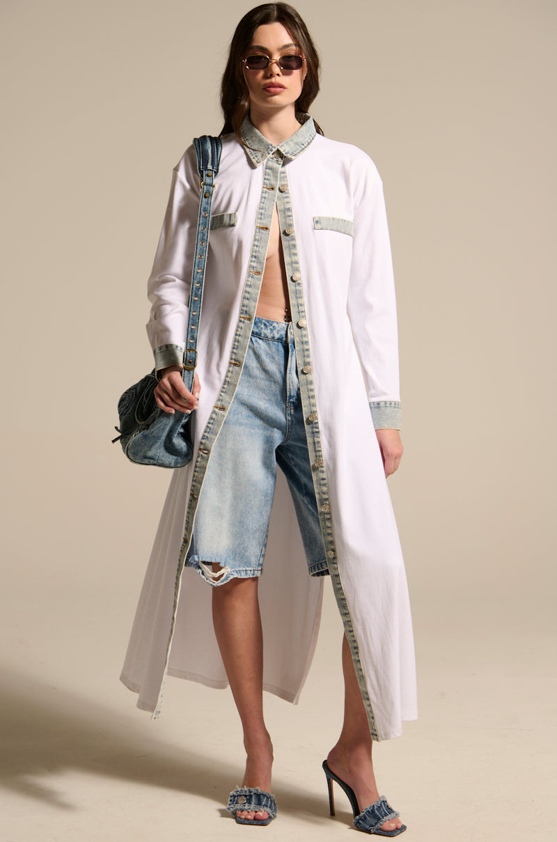 Wearing sunglasses, the woman stands against a plain background styled in the LEILA OVERSIZED WHITE MULTI BUTTON UP, distressed knee-length denim shorts, denim high heels, and a blue quilted shoulder bag.