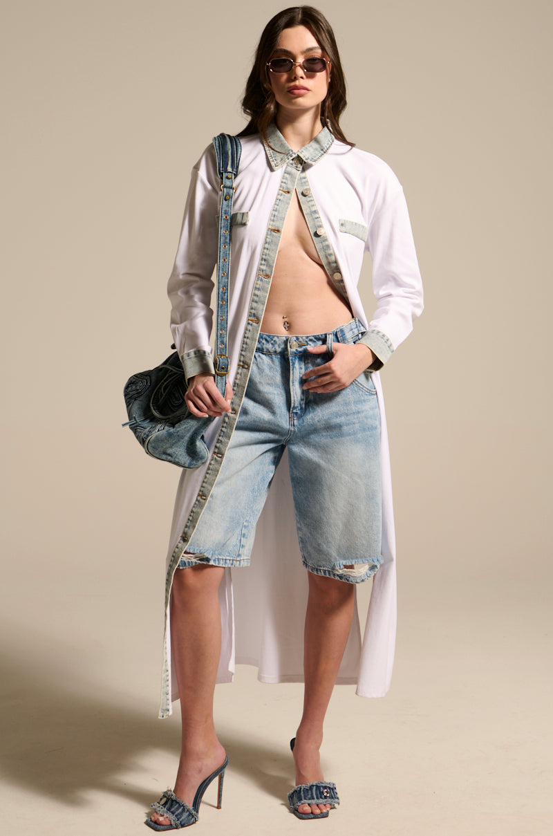 A woman wearing sunglasses, heeled sandals, denim Bermuda shorts, and carrying a denim bag stands confidently against a plain background in the LEILA OVERSIZED WHITE MULTI BUTTON UP with light-washed denim trim.