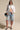 A woman wearing sunglasses, heeled sandals, denim Bermuda shorts, and carrying a denim bag stands confidently against a plain background in the LEILA OVERSIZED WHITE MULTI BUTTON UP with light-washed denim trim.