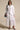 A woman wearing the LEILA OVERSIZED WHITE MULTI BUTTON UP with light-wash denim trim, sunglasses, a denim shoulder bag, and blue open-toe heels stands against a beige background.