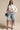 A woman stands confidently against a plain backdrop wearing sunglasses, knee-length light-wash denim shorts, blue heels, a blue handbag, and the LEILA OVERSIZED WHITE MULTI BUTTON UP shirt left mostly unbuttoned.