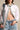A woman wears the LEILA OVERSIZED WHITE MULTI BUTTON UP with blue jeans, revealing her midriff and a navel piercing. A denim bag strap rests over her shoulder.