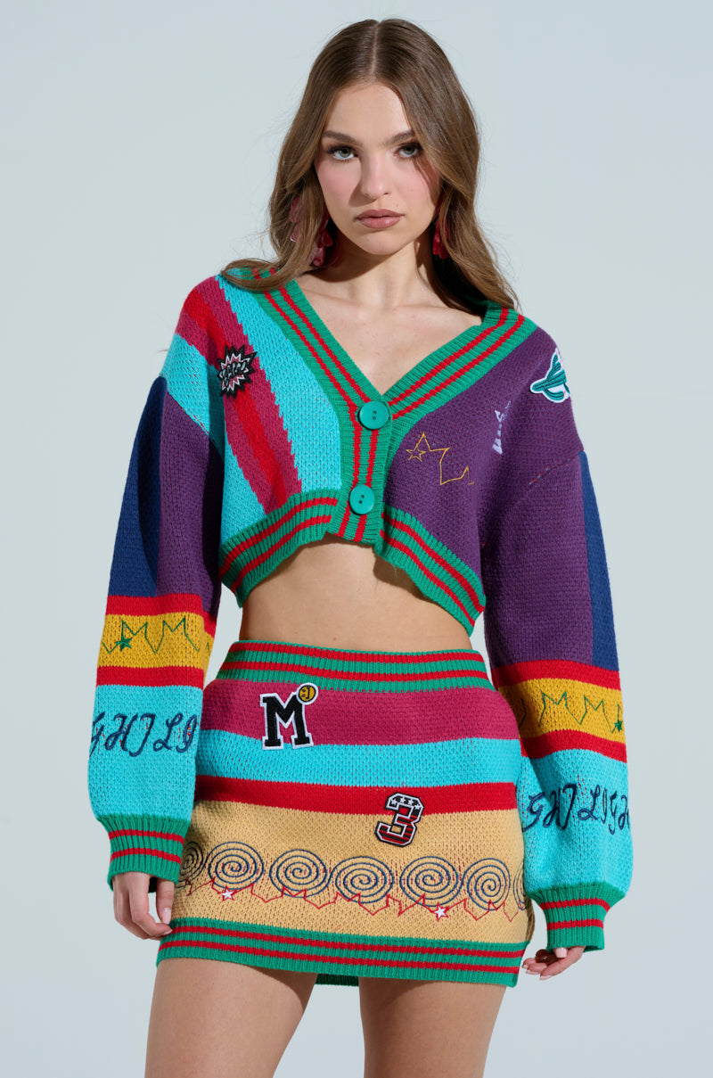 A woman proudly displays the "Free Falling Cardigan," a vibrant color-block ensemble featuring a patterned knit with stripes, embroidered symbols, and text. The cropped cardigan pairs with a matching mini skirt, her long hair and large earrings beautifully complement the light background.