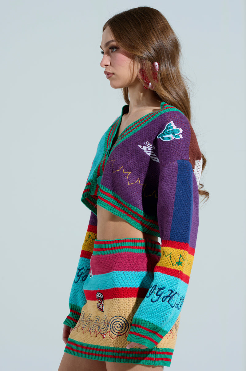 A woman with long brown hair in profile wears the FREE FALLING CARDIGAN, a colorful knit outfit with an oversized cropped sweater and matching skirt. The ensemble is color-blocked with teal, red, purple, and yellow patterns. She completes her look with pink drop earrings.