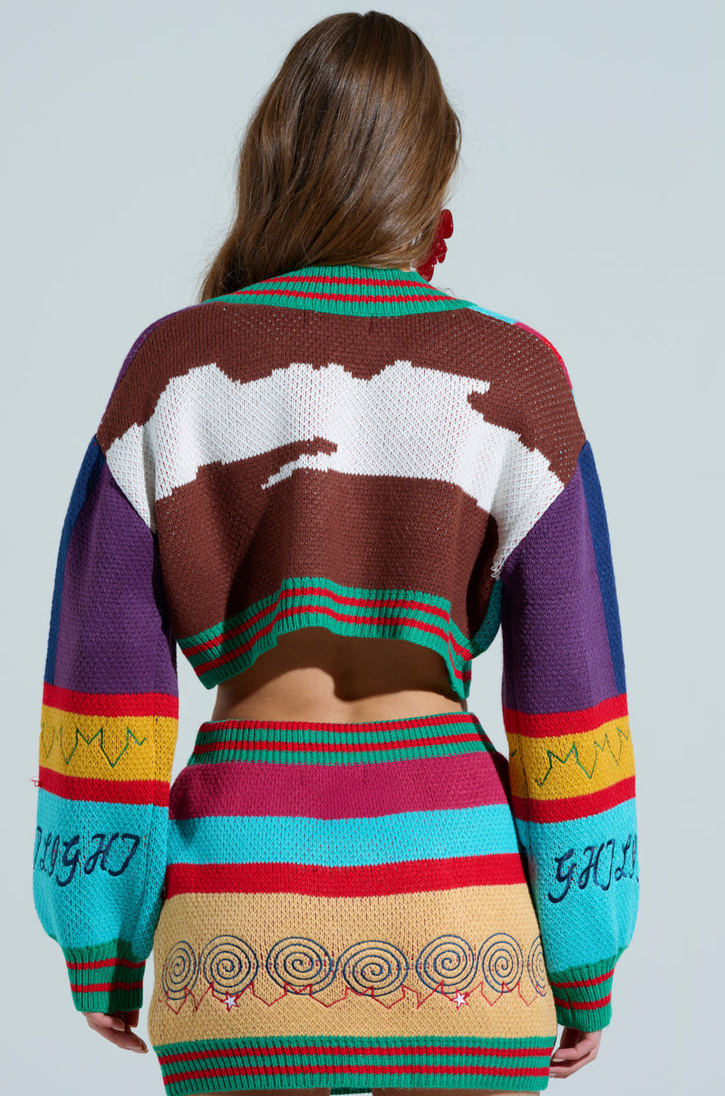 A person with long brown hair wears a colorful knitted FREE FALLING CARDIGAN showcasing abstract patterns in brown, white, and blue on the back. The outfit includes a skirt with horizontal stripes and spiral designs alongside lettered sleeves against a light gray background.