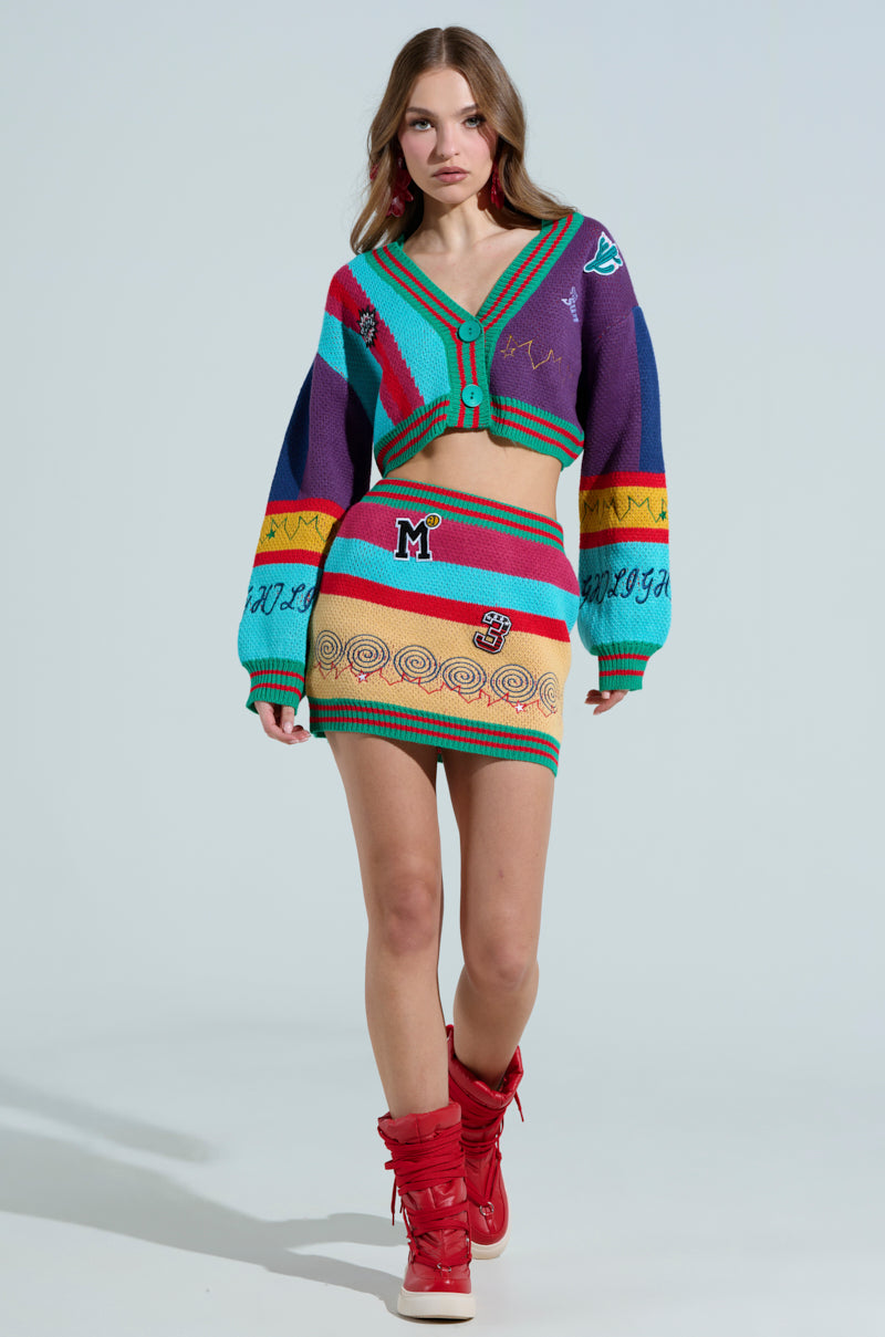 The woman is wearing the FREE FALLING CARDIGAN, a colorful knitted two-piece outfit with bold vibrant stripes, a cropped cardigan, and mini skirt. She pairs it with red high-top boots while walking toward the camera against a plain background.