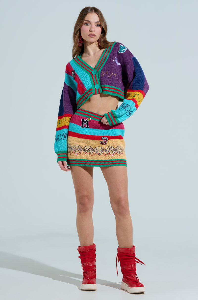 A woman stands wearing a vibrant FREE FALLING CARDIGAN and skirt set with assorted patterns and letters. She has red boots, her hands by her sides, against a plain light blue backdrop.