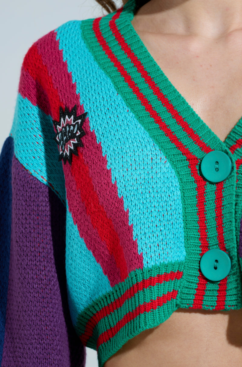 A close-up features a person donning the FREE FALLING CARDIGAN, a vibrant knit with striped patterns in teal, red, and purple. This oversized sweater has large teal buttons and a comic-style patch on the front. The person's face is not visible.