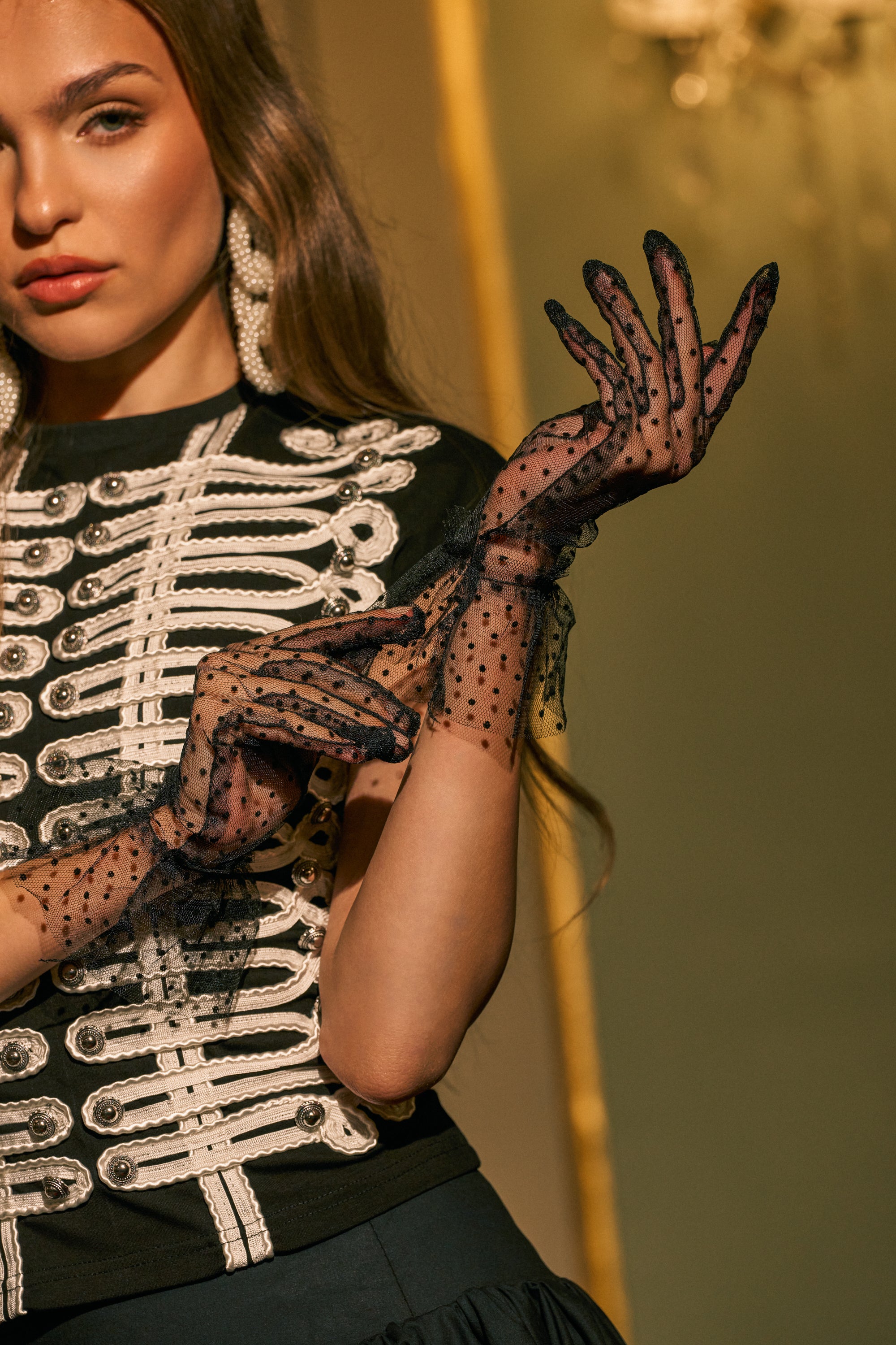 A woman wearing a decorative black and white jacket, statement earrings, and the OH YOU FANCY DOTTED GLOVES poses with one hand adjusting her glove. Only the upper half of her body is visible.