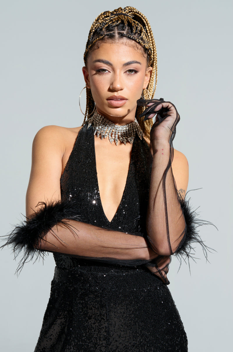 A person with braided hair wears a glamorous black sequin dress and Featherington Gloves, which feature exquisite feather trim detailing. A sparkling choker necklace enhances the elegance as they pose confidently against a light background, holding part of the Featherington Glove near their face.