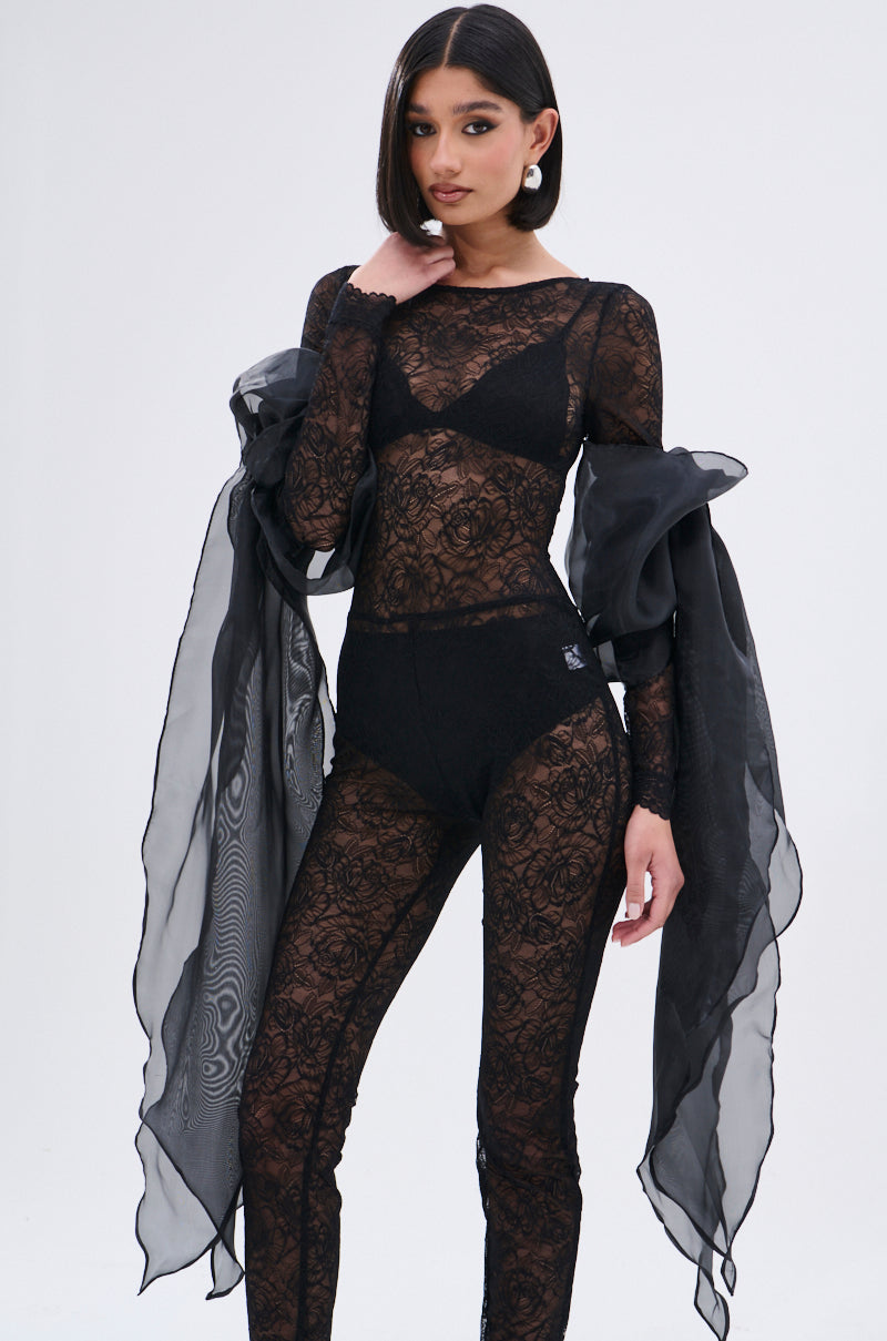 A woman poses in sheer black lace with oversized puff shoulders, high-waisted shorts, and the OVER THA TOP SLEEVES—sheer flowing black organza draped over her arms—paired with a black bralette and large pearl earrings.