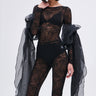 A woman poses in sheer black lace with oversized puff shoulders, high-waisted shorts, and the OVER THA TOP SLEEVES—sheer flowing black organza draped over her arms—paired with a black bralette and large pearl earrings.
