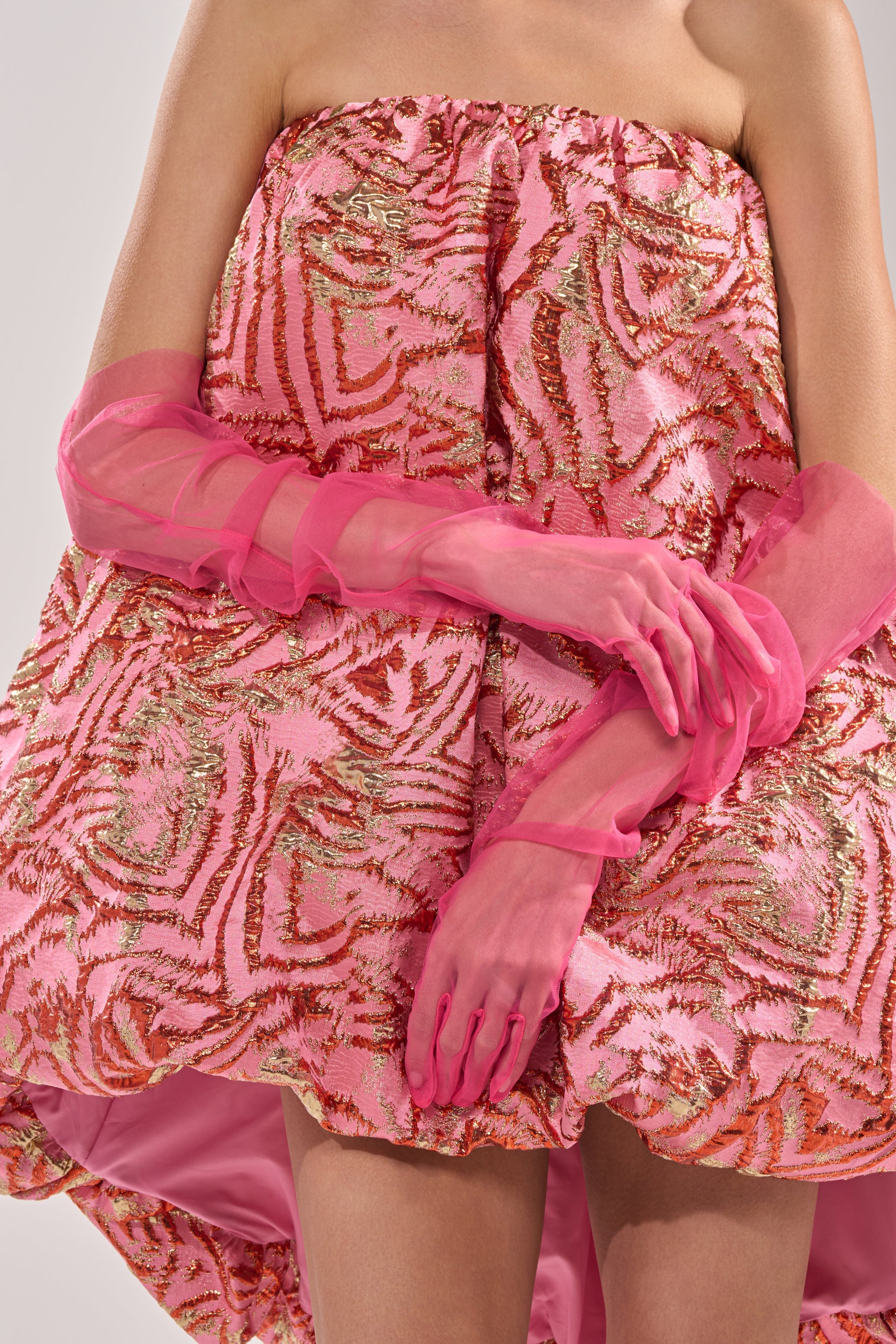 A person stands against a neutral background wearing voluminous, strapless pink and gold-patterned dress and bright pink DOLL MESH GLOVES on their upper arms; the upper face is not visible. 0