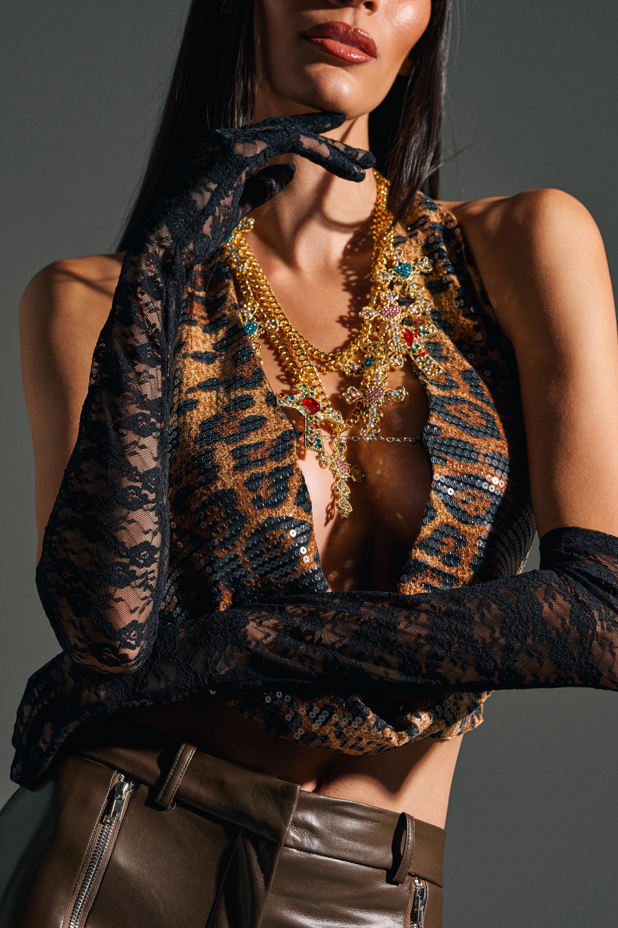 A woman poses against a dark background in UNTIL THE MORNING LACE GLOVES, pairing the sheer black gloves with a sequined leopard-print top, layered gold charm necklaces, and brown pants, one hand gracefully near her face. 2