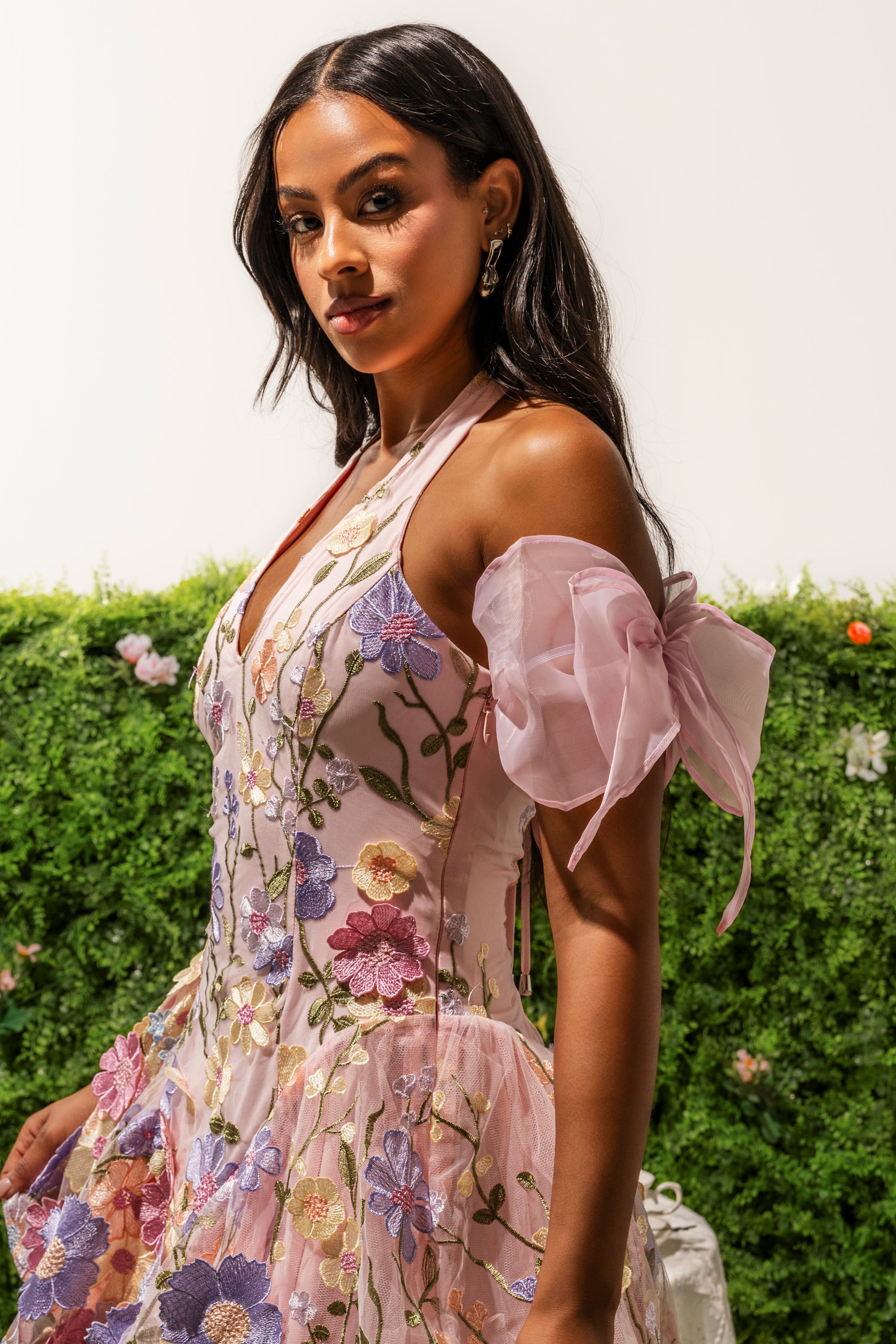 A woman with long dark hair wears the "TIE IT WITH A BOW SLEEVES" sleeveless pink dress, featuring floral embroidery and sheer ruffled off-shoulder sleeves, standing before a green leafy background with small flowers. 0