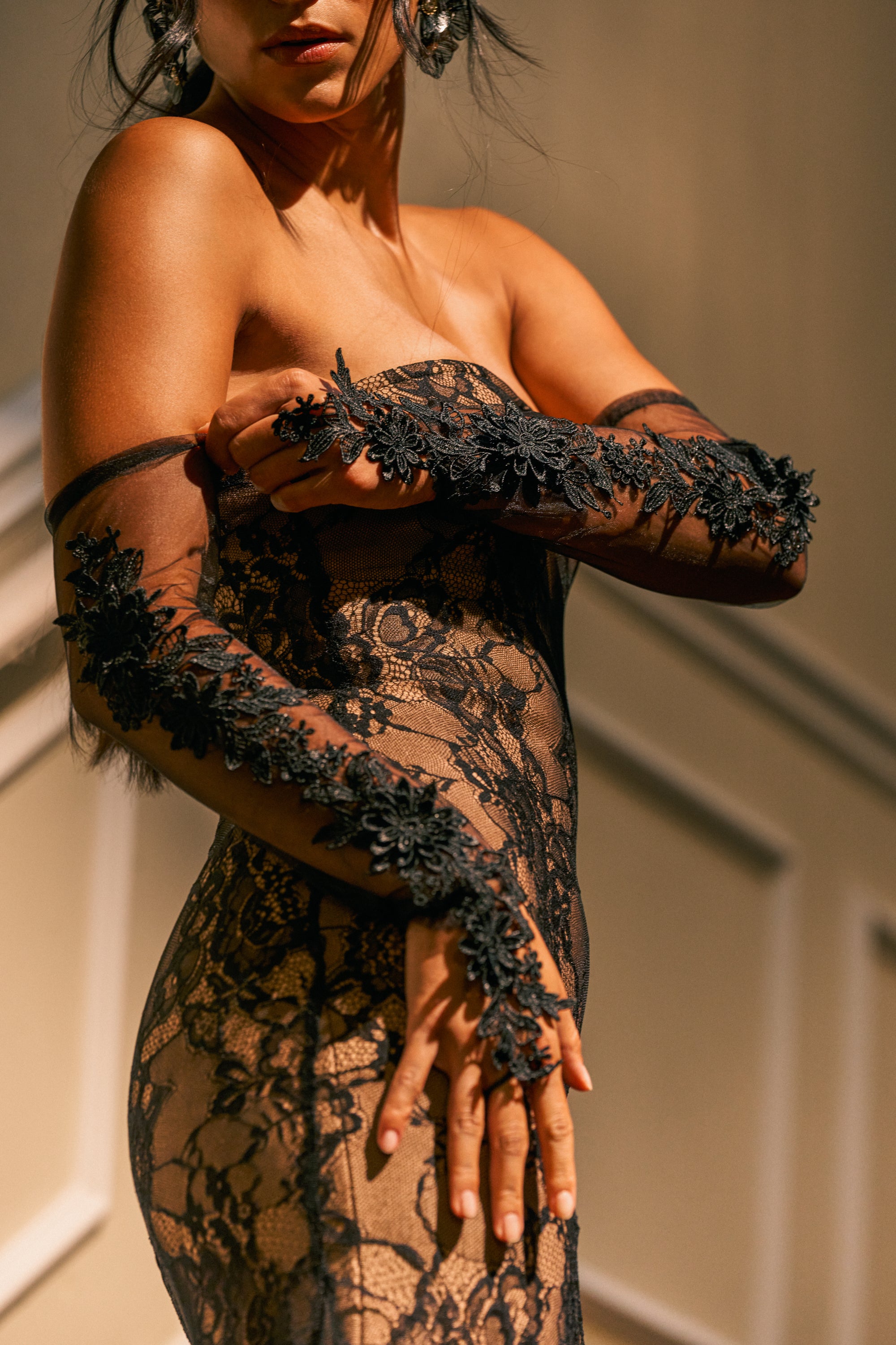 Wearing the IM SO FANCY APPLIQUE FINGERLESS GLOVES, a woman poses indoors with her hands gracefully positioned, partially covering her chest, as she dons a strapless sheer black lace dress adorned with intricate floral details.
