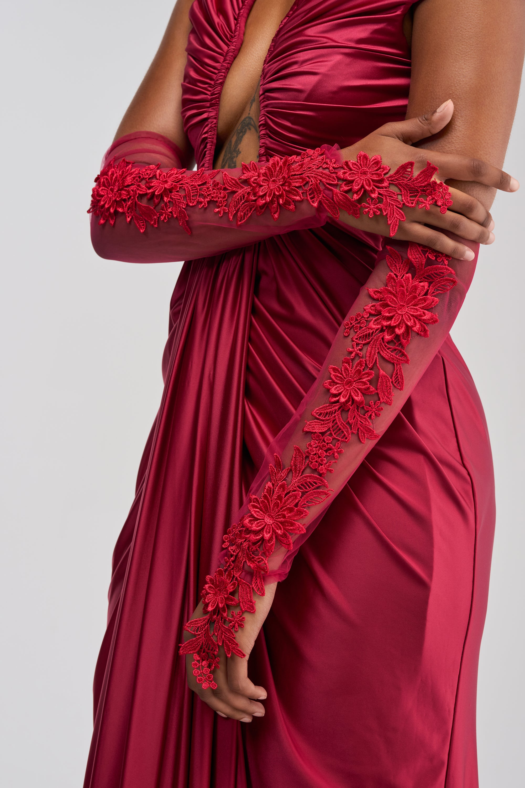 Woman in a red satin dress with arms crossed, sporting the ULTIMATE APPLIQUE FINGERLESS GLOVES IN BURGUNDY—burgundy mesh gloves adorned with red floral embroidery—showing off a tattoo on her upper arm.