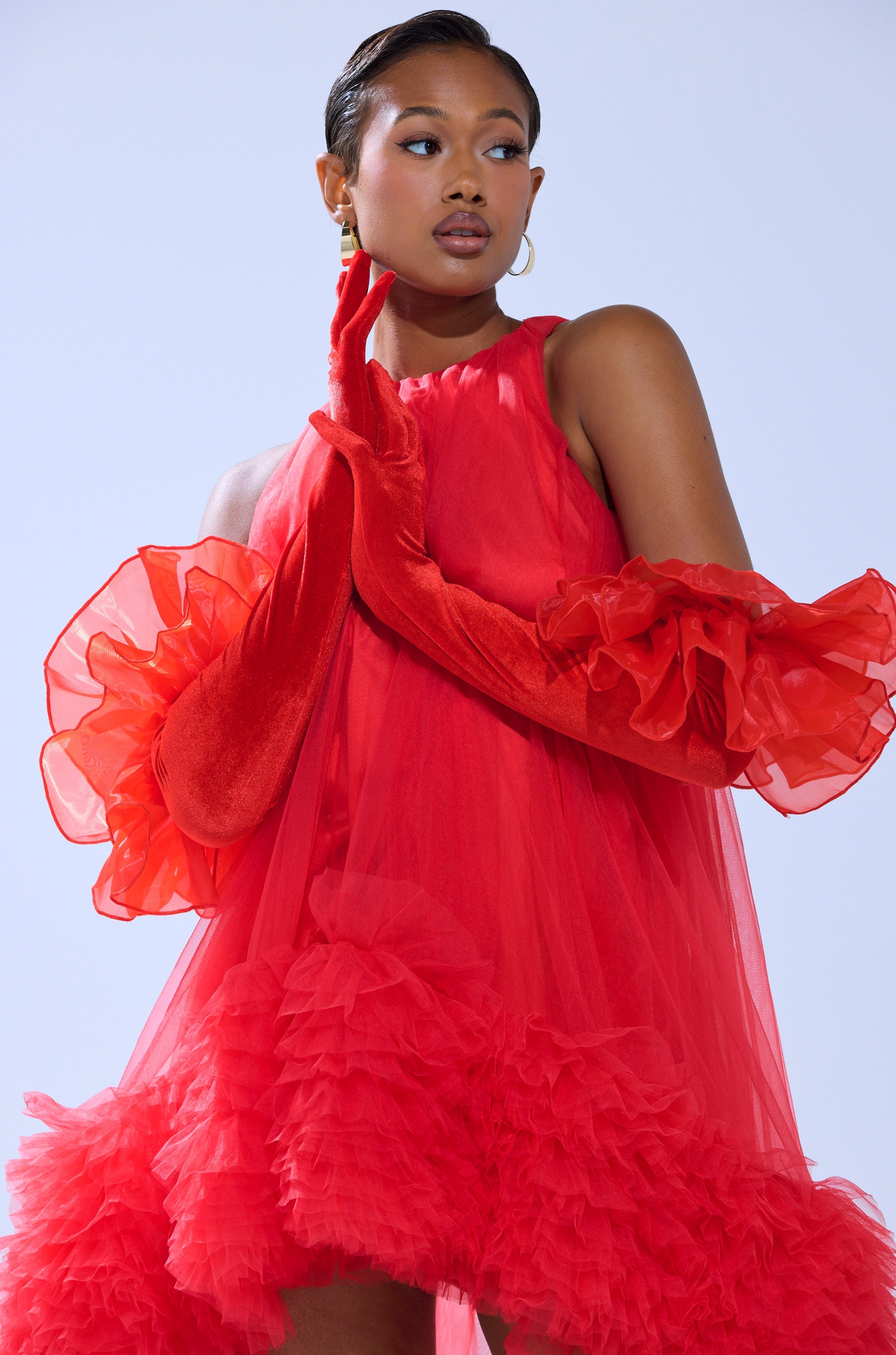 A woman poses confidently against a light blue background, wearing dramatic AT THE GALA VELVET GLOVES with a vibrant red, ruffled dress crafted from sheer, flowy fabric and accented by voluminous details. 0