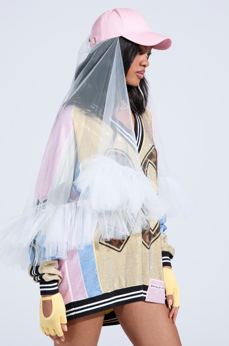 A person wearing an AKIRA Label multicolored jacket with tulle and mesh details. Their CITY NIGHTS VEILED HAT, a pink cap with a veil partially covering their face, is stylishly paired with yellow fingerless gloves. The jacket stands out with bold patterns and a unique layered design. 0