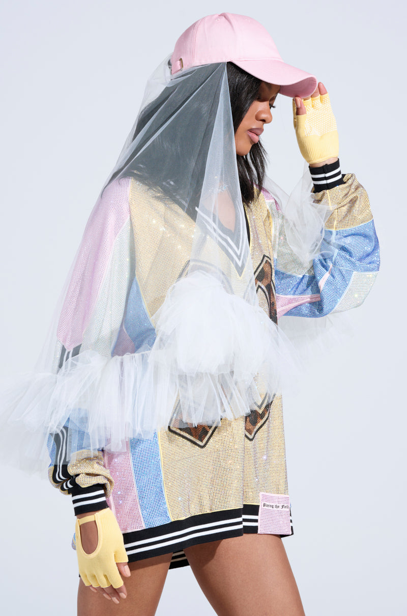 The model flaunts a vibrant oversized shirt with sequins, stripes, and tulle details. Completing the ensemble is the CITY NIGHTS VEILED HAT—an AKIRA Label baseball cap with a sheer veil—paired flawlessly with yellow fingerless gloves against a plain light gray backdrop. 1