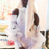 A person stands in a convenience store wearing the TAYTAY TULLE SLEEVES, a sequined white dress featuring sheer, tulle sleeves. They complete their look with a fluffy white hat adorned with long ear flaps. Bottled drinks are visible in the refrigerated display behind them, enhancing the scene's dramatic silhouette.