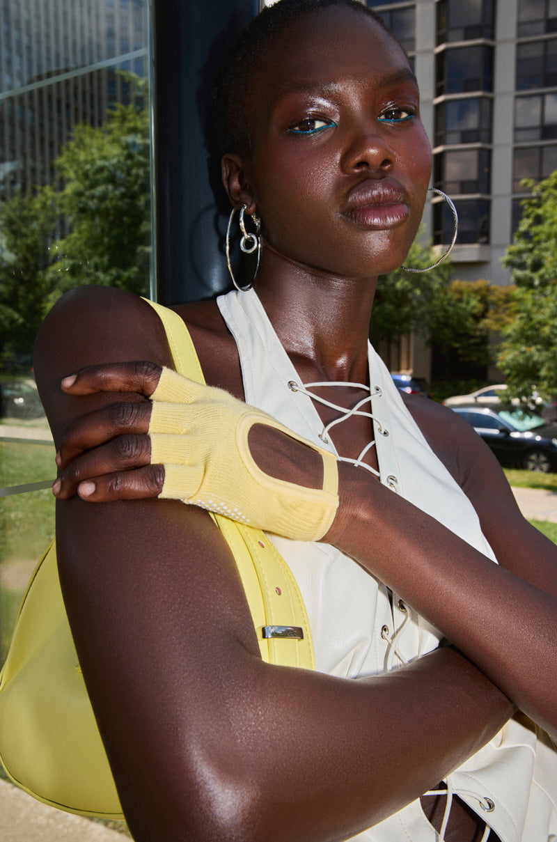 A person stands outdoors with arms crossed, wearing a sleeveless lace-up top, silver hoop earrings, and trendy TOO TRENDY GLOVES knit fingerless gloves, holding a large yellow bag. Green trees and city buildings are visible in the background.