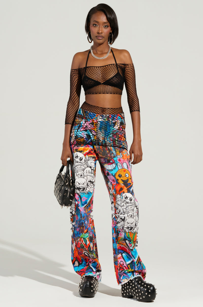A person stands confidently wearing a black fishnet crop top and vibrant SAMMIE CARTOON DENIM JEANS. They hold a studded black bag and rock platform shoes adorned with studs, all set against a plain off-white background.